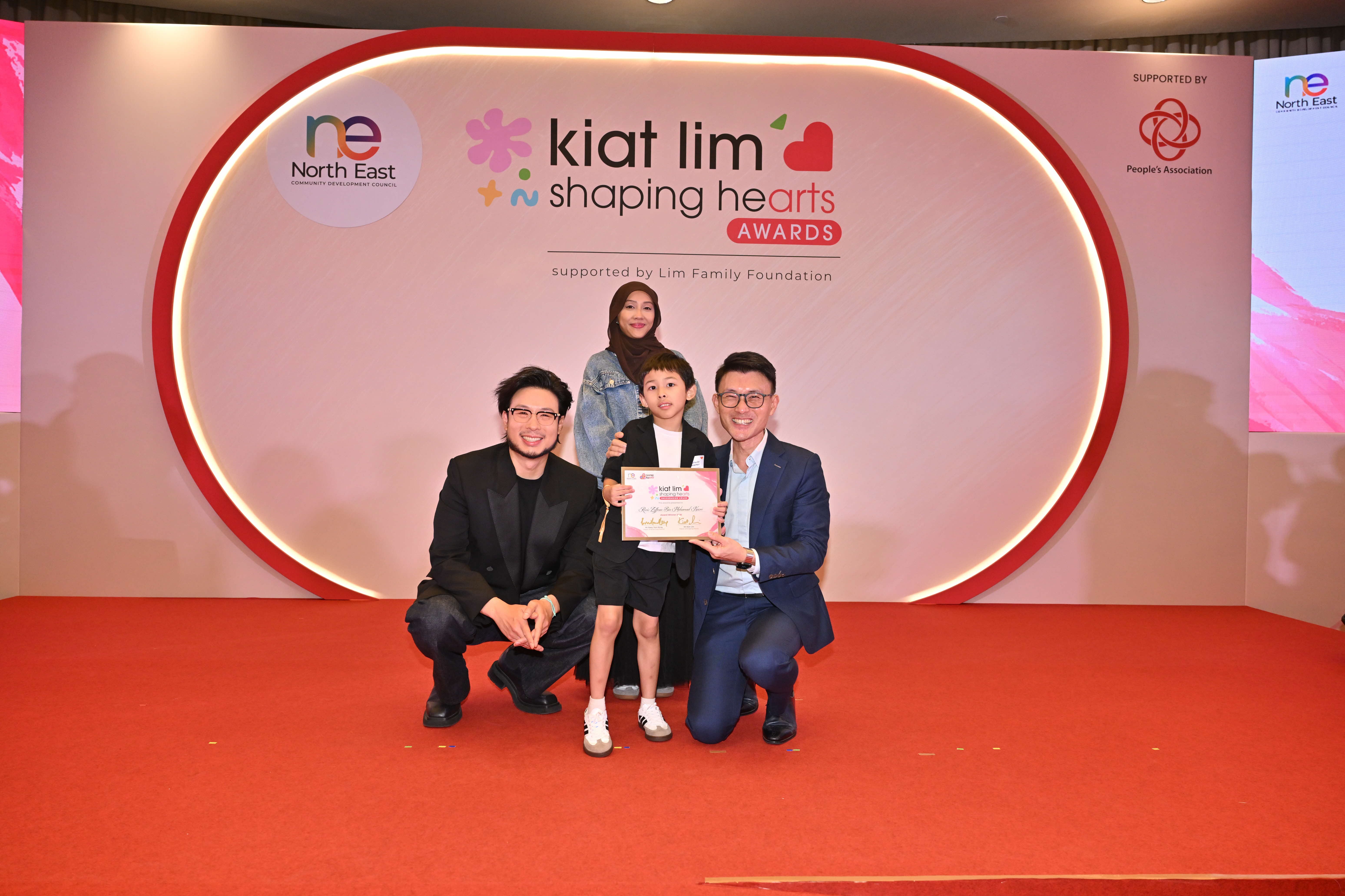 Award recipient standing on stage with the presenter, holding their certificate and posing for a group photo during the award ceremony, with a backdrop reading ‘Kiat Lim-Shaping Hearts Awards'.